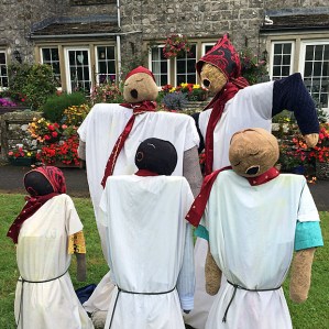 Five scarecrows singing in front of a colourful garden