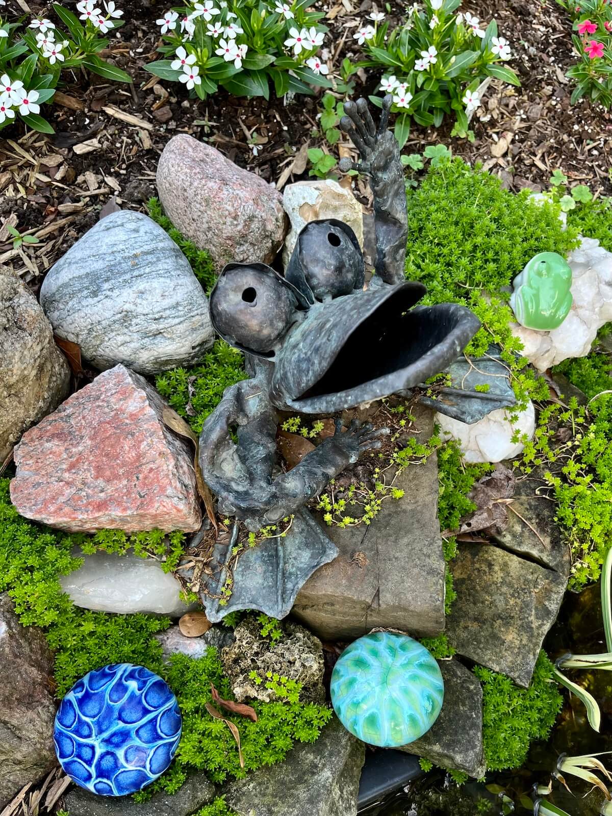 Wide-mouthed frog fountain surrounded by stones, glass orbs, moss and flowers